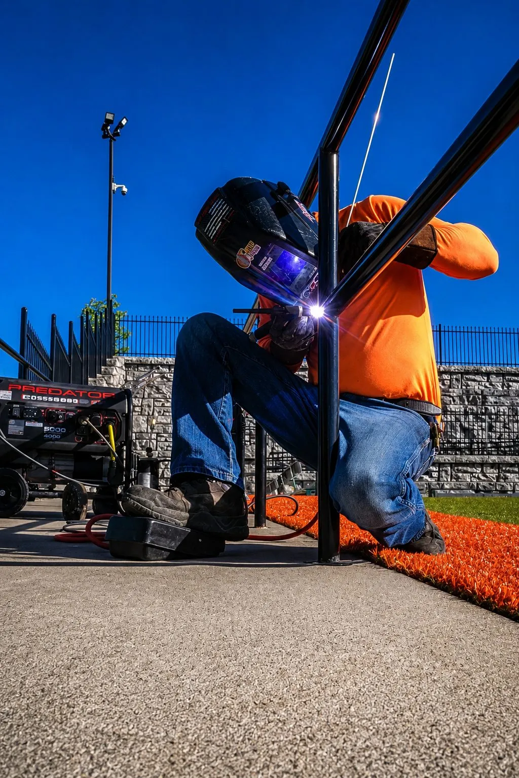 Tyler kneeling on a rubber running track, welding a black steel rail post at a ballfield fence under a clear blue sky.