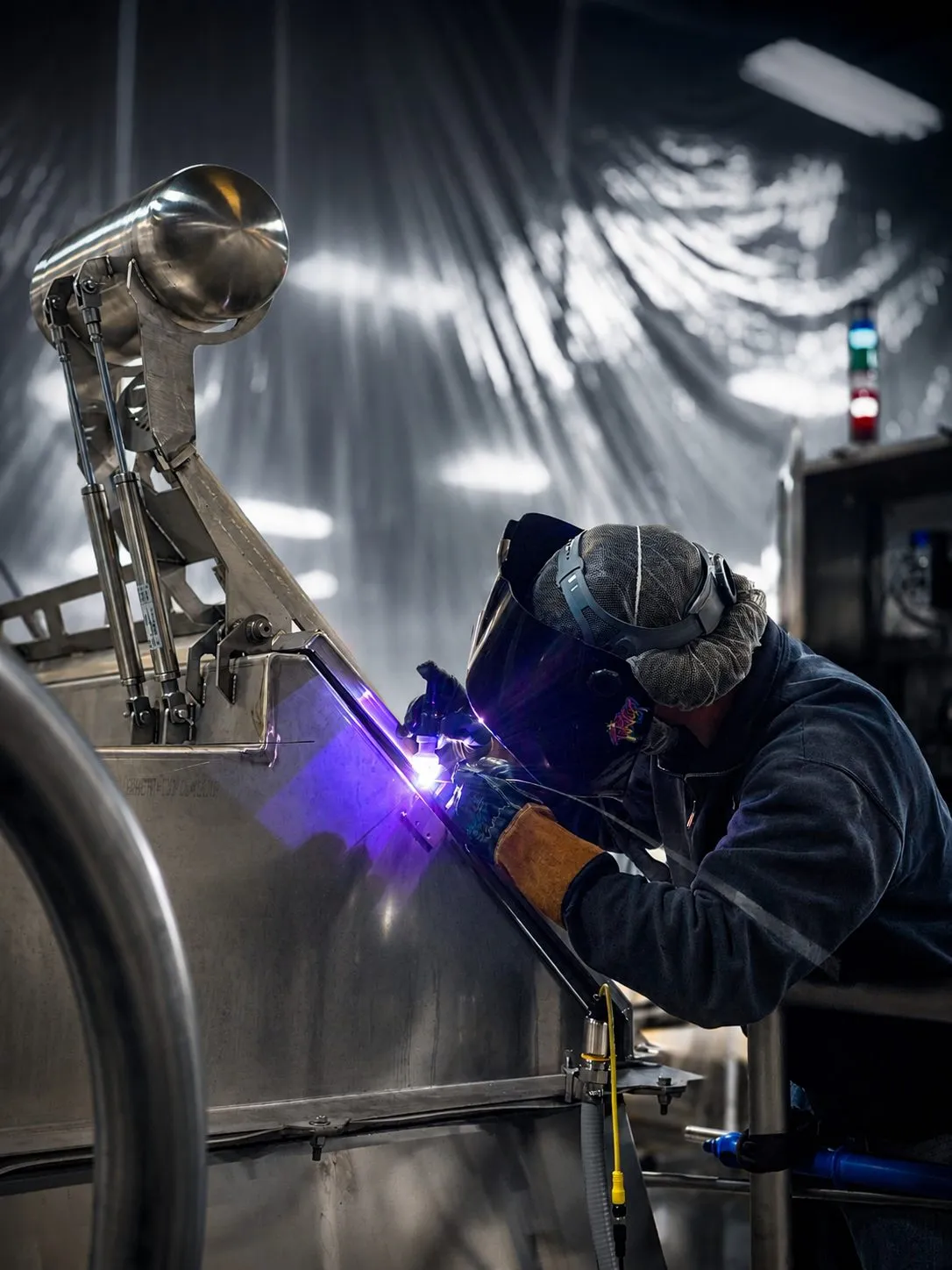 Tyler TIG welding stainless steel on a brewery tank behind plastic sheeting, the arc lighting the fit-up in blue.