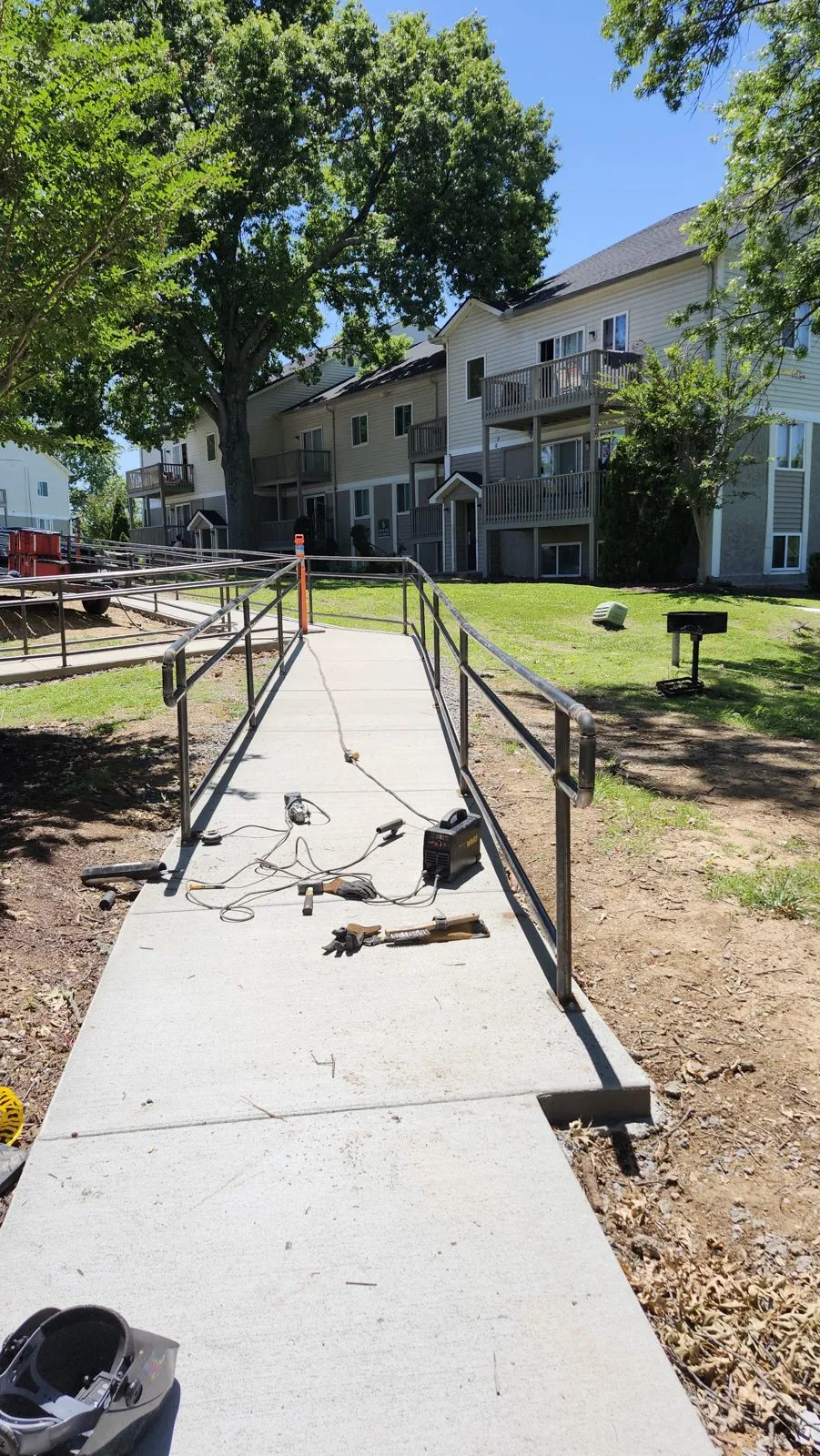 A long run of black steel handrail along a concrete sidewalk ramp between apartment buildings, welding leads still on the ground.