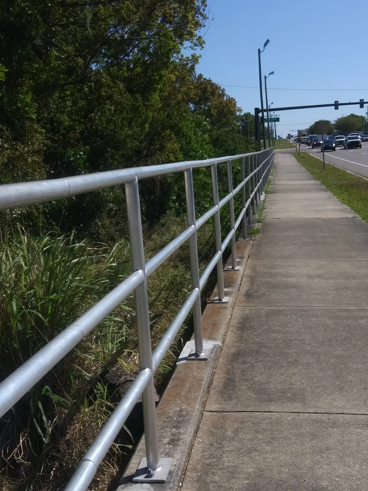 Galvanized pipe handrail on posts running the length of a concrete sidewalk beside a road.