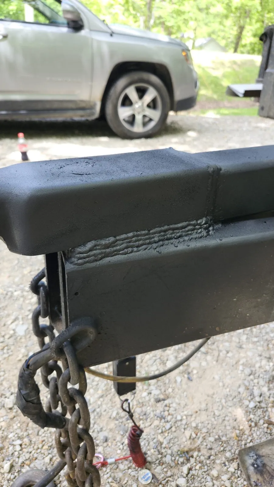 A trailer tongue and coupler with a fresh repair weld primed black, on a gravel drive with the tow vehicle behind it.