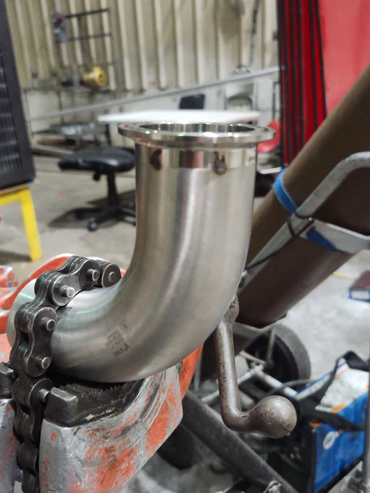 A polished stainless elbow with a tri-clamp ferrule held in a pipe vise, ready to be fit to the line.