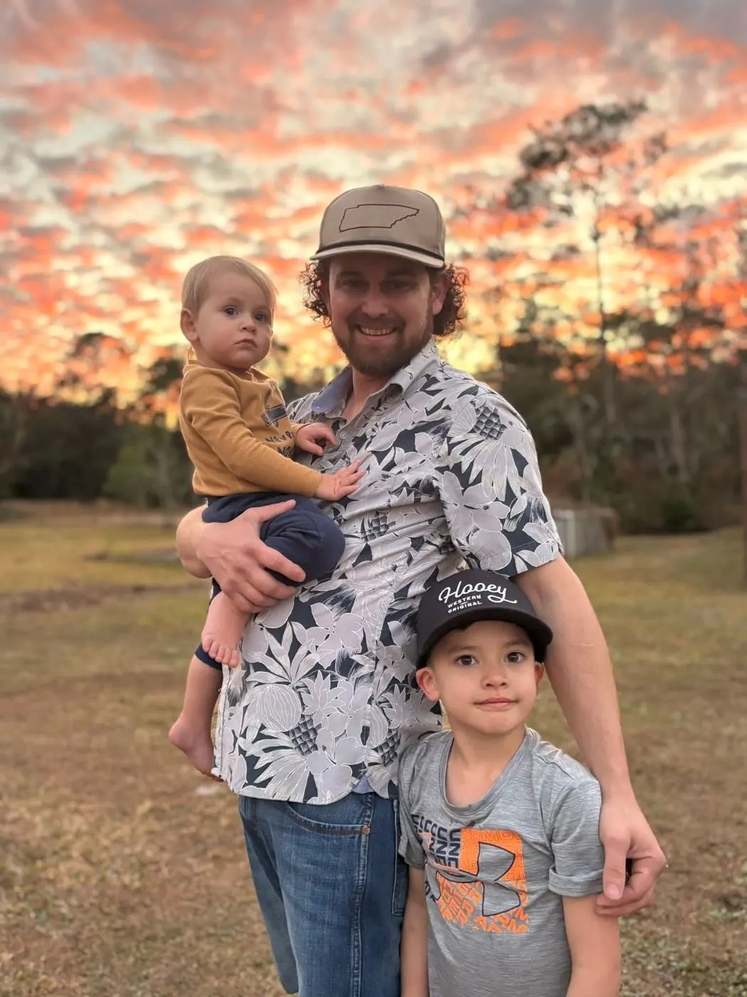 Tyler Edwards holding his two young children in a field under a sunset sky